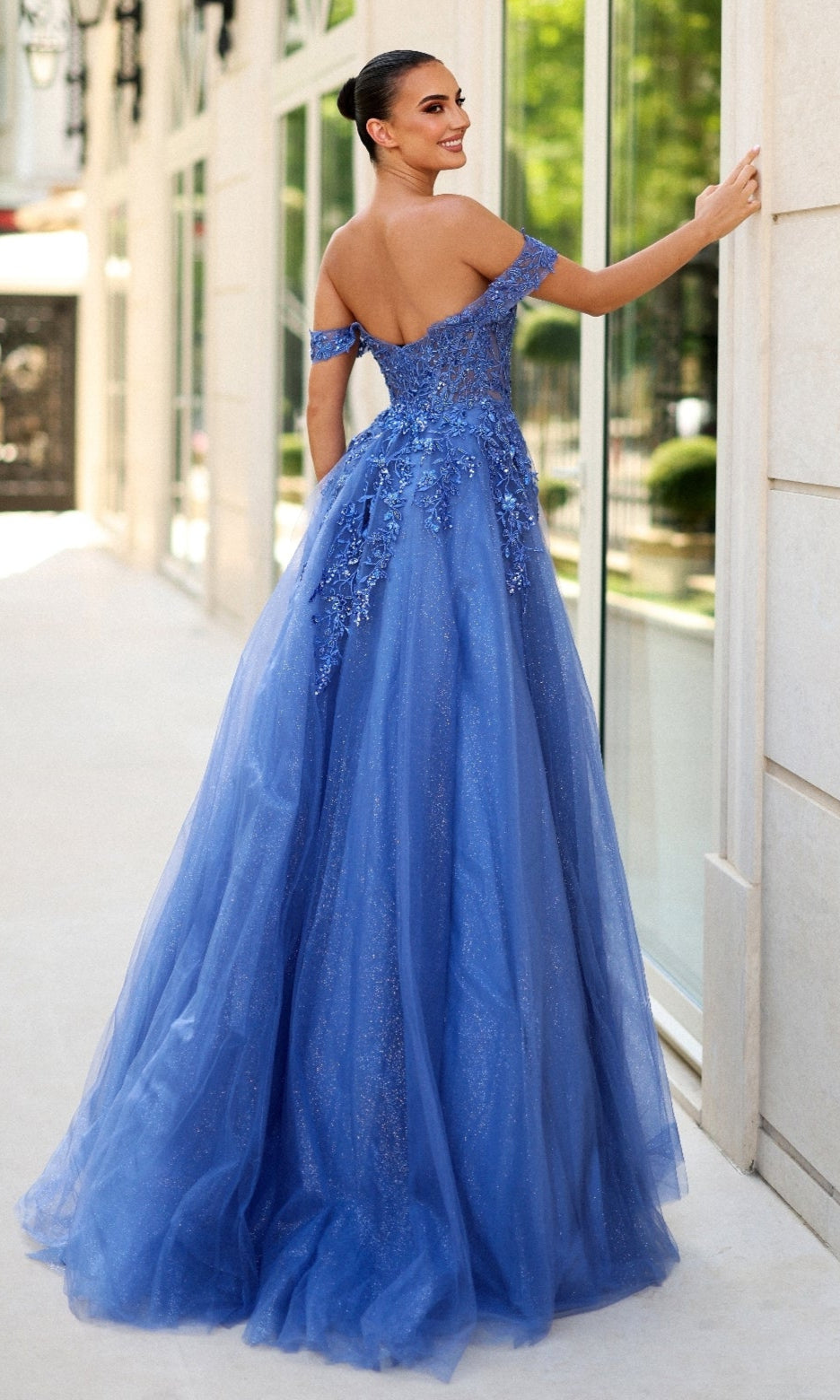 A woman in the Long Glitter Blue Prom Dress: Tina Holly TE232 with floral embellishments, an off-the-shoulder corset bodice, and a glitter tulle skirt stands outdoors, smiling over her shoulder.